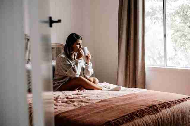 a woman drinking her coffee in bed while looking out the window 